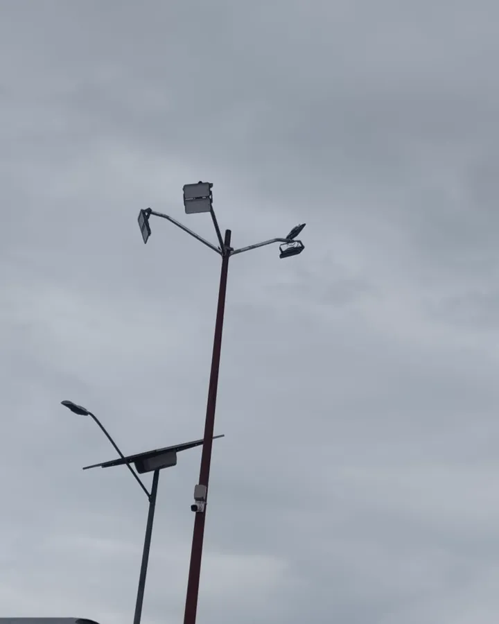 Poste con reflectores bajo un cielo nublado y, a su lado, otro poste con una luminaria y un panel solar